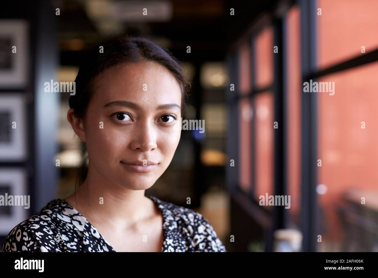 Tausendjährige asiatischen Geschäftsfrau durch Fenster im Büro zu Kamera, Kopf und Schultern suchen, Nahaufnahme Stockfoto
