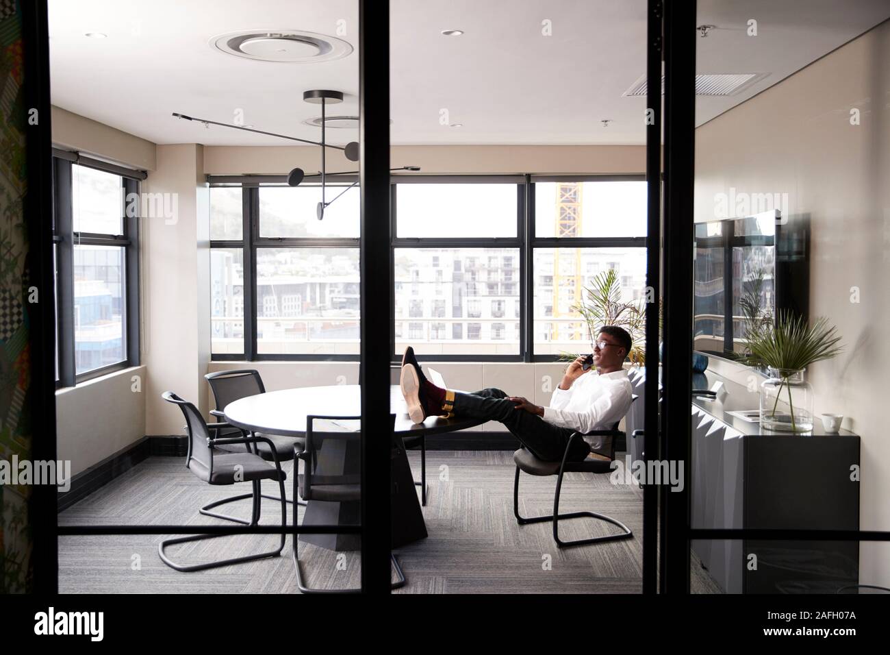 Tausendjährige schwarzer Geschäftsmann am Telefon zu sprechen mit den Füßen auf einem Schreibtisch, vom Eingang gesehen Stockfoto