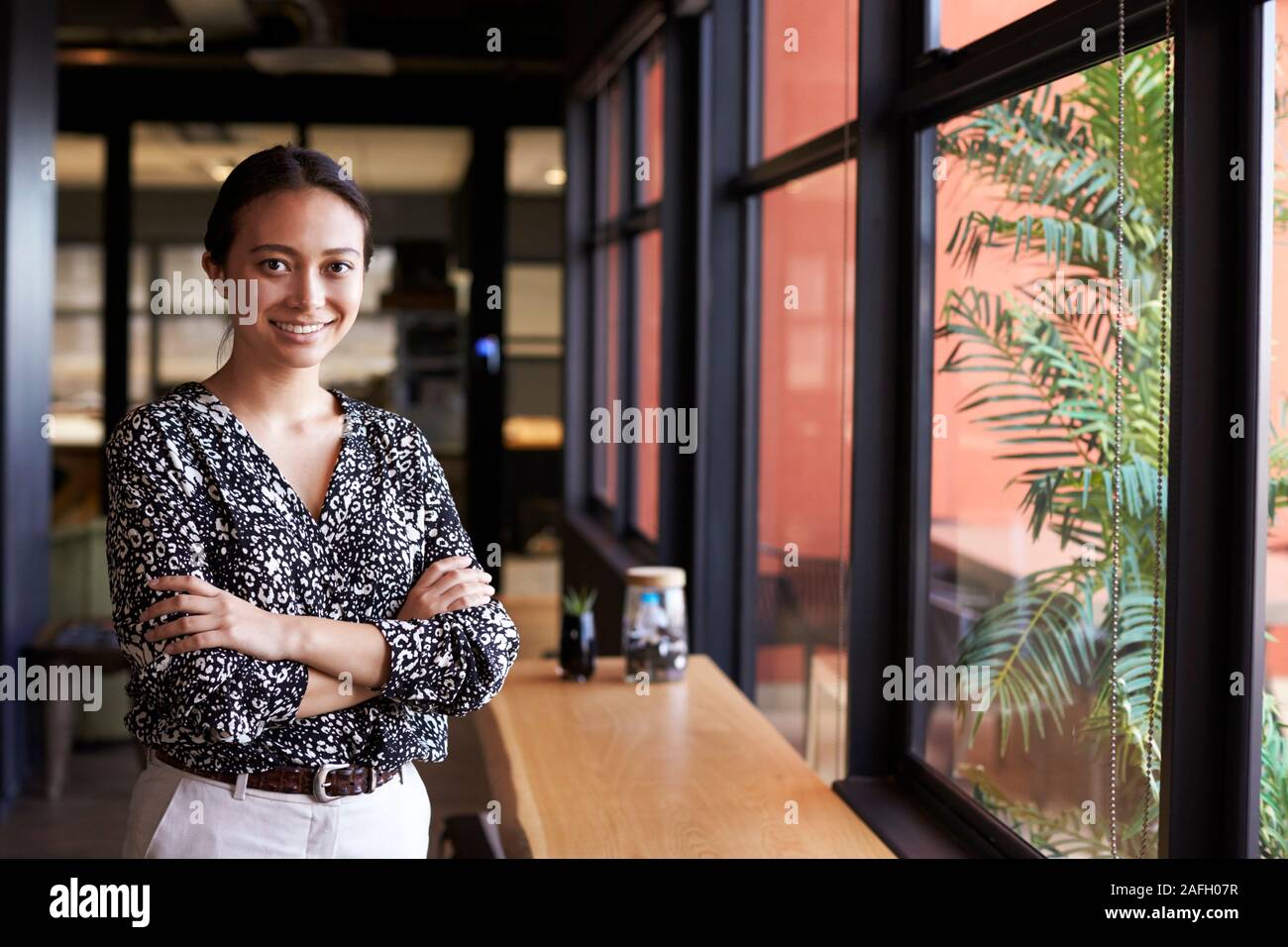 Tausendjährige asiatischen Geschäftsfrau stehen durch das Fenster in ein Büro in die Kamera lächelt, Taille bis Stockfoto