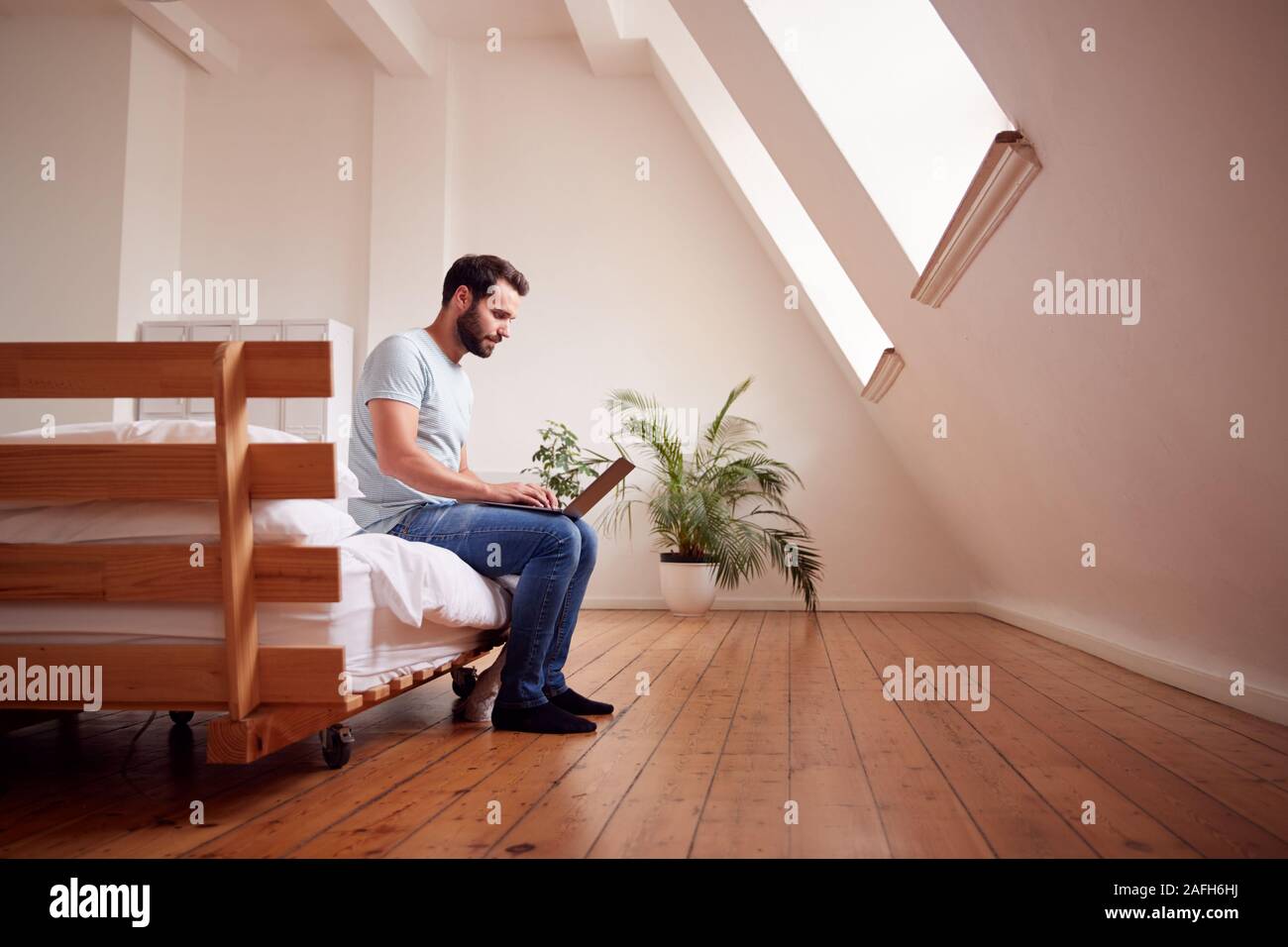 Junge Mann sitzt auf Bett im Loft Apartment Arbeiten am Laptop Stockfoto