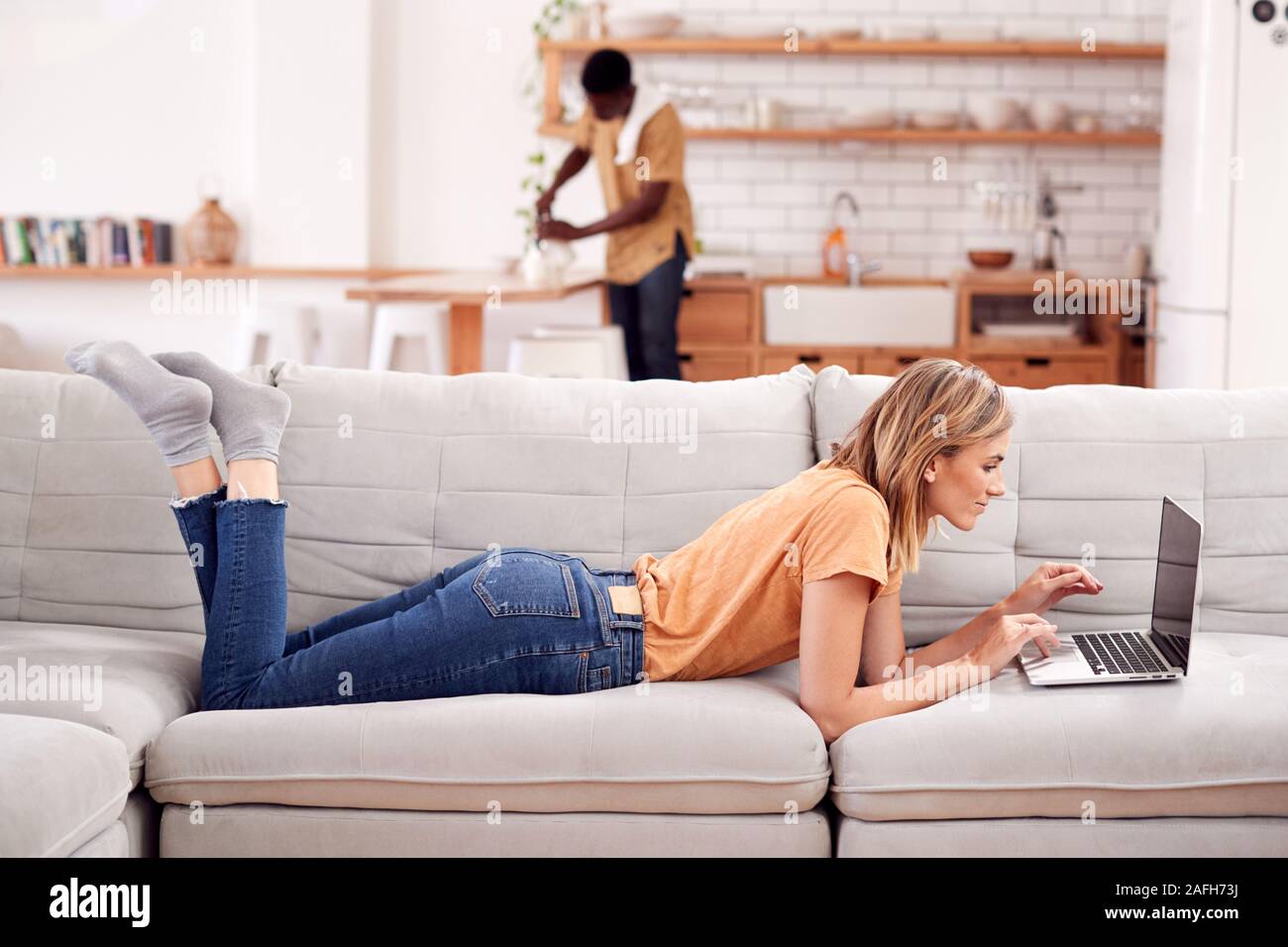 Frau Entspannende liegend auf dem Sofa zu Hause mit Laptop mit Mann in der Küche hinter Stockfoto