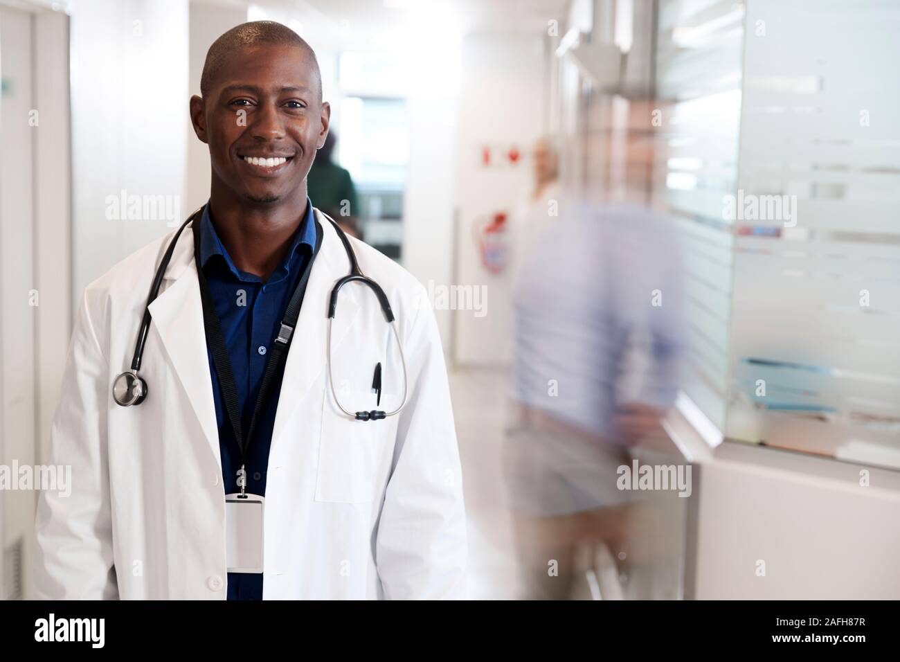 Portrait von lächelnden männlicher Arzt tragen weiße Mantel mit Stethoskop in Langen Krankenhaus Flur Stockfoto