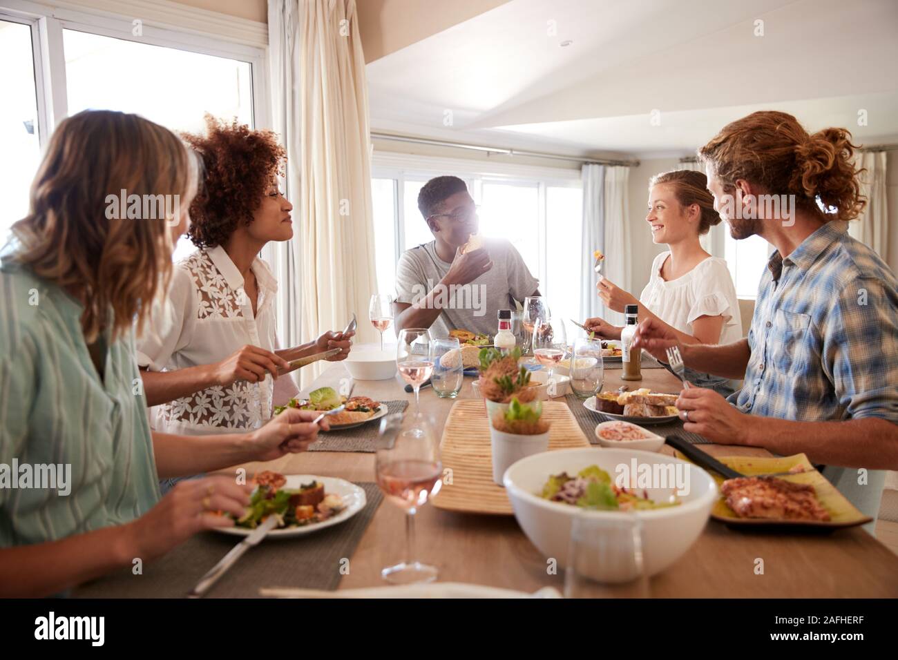 Eine Gruppe von tausendjährigen Freunden an einem Tisch sitzen, essen und reden, mit Hintergrundbeleuchtung Stockfoto