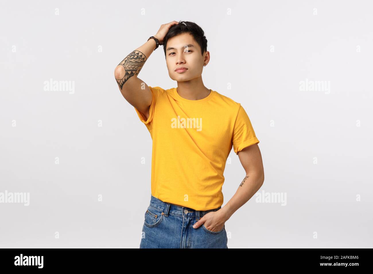 Stilvoll und sicher gut aussehende Chinesische tätowierten Mann im gelben T-Shirt, den Fingern durch die Haare und Suchen selbstbewusste Kamera, selbst Liebe und Stockfoto