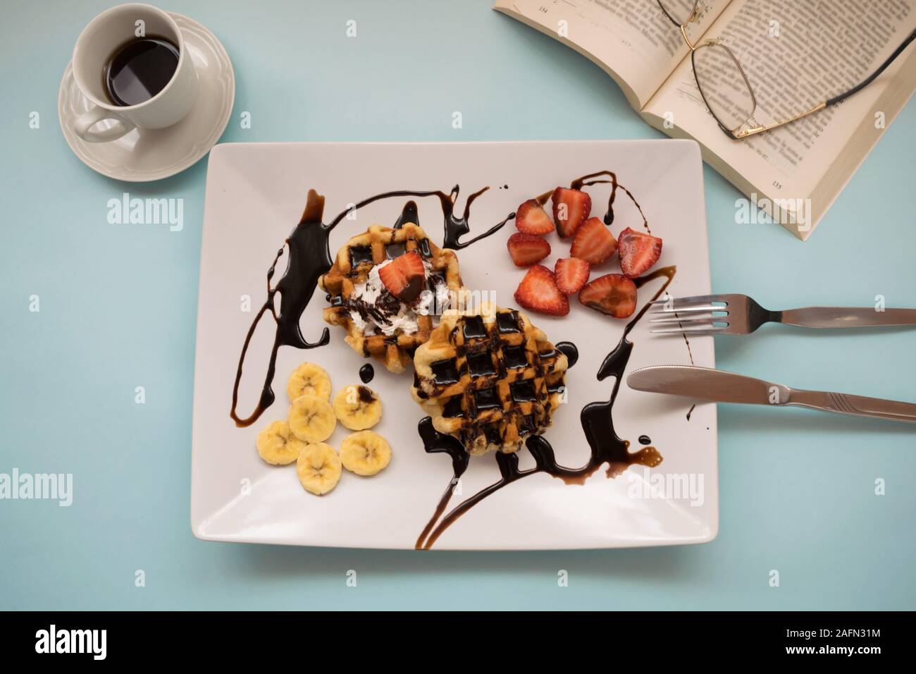 Waffeln mit Schokolade, Obst, eine Tasse Kaffee und ein Buch auf einem blauen Hintergrund Stockfoto
