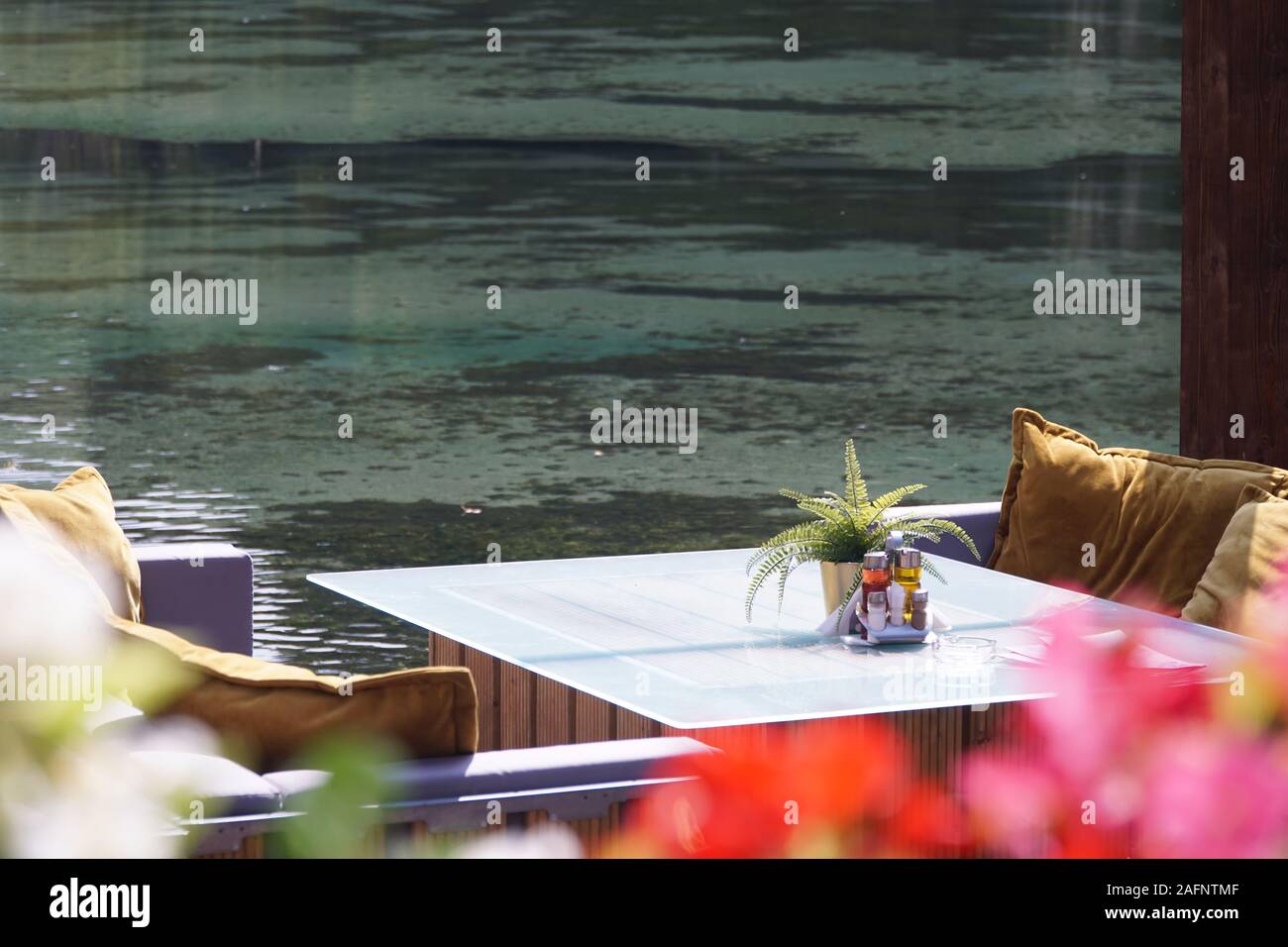 Ein Glas Tisch in einem Restaurant mit Blick auf den See oder Fluss mit Dekorationen, eine Reihe von Gewürzen, einen Topf mit einer Pflanze. Ein sonniger Sommertag. Niemand. Stockfoto