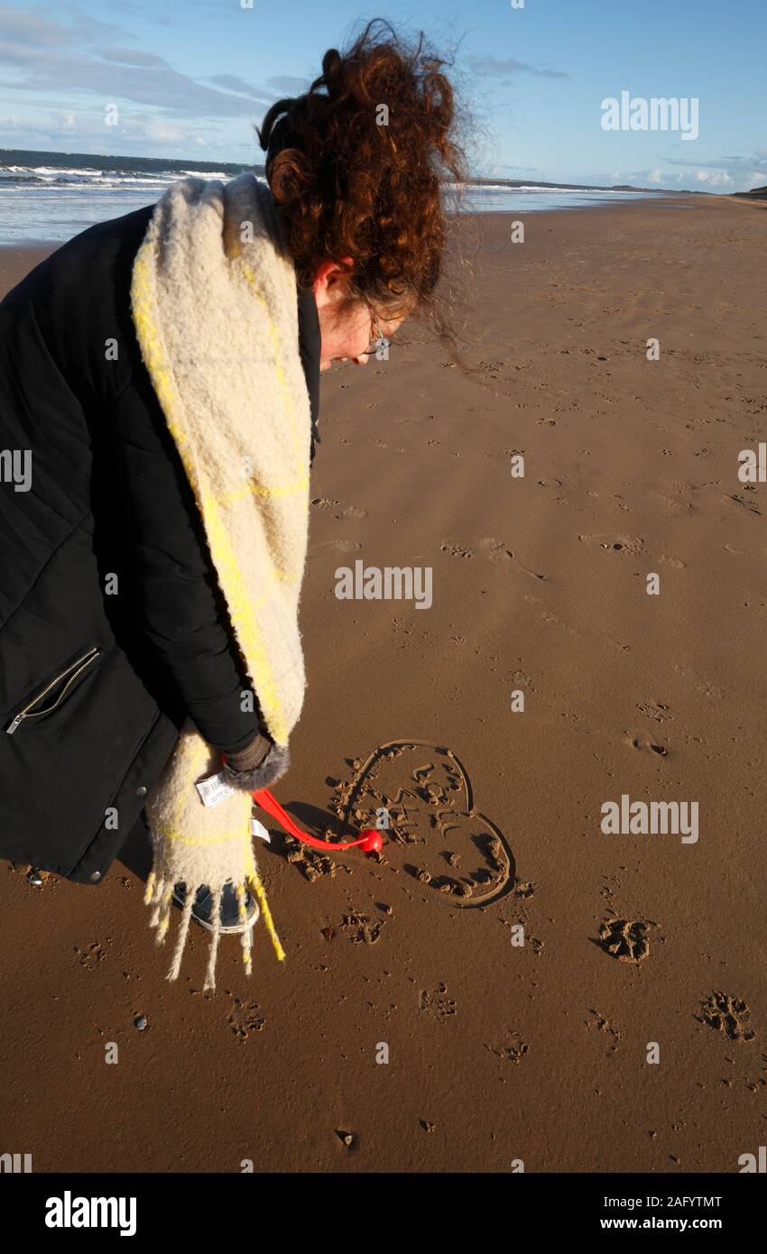 Frau, die ein Liebesherz im Sand bei Brancaster, Norfolk, zeichnet. Stockfoto