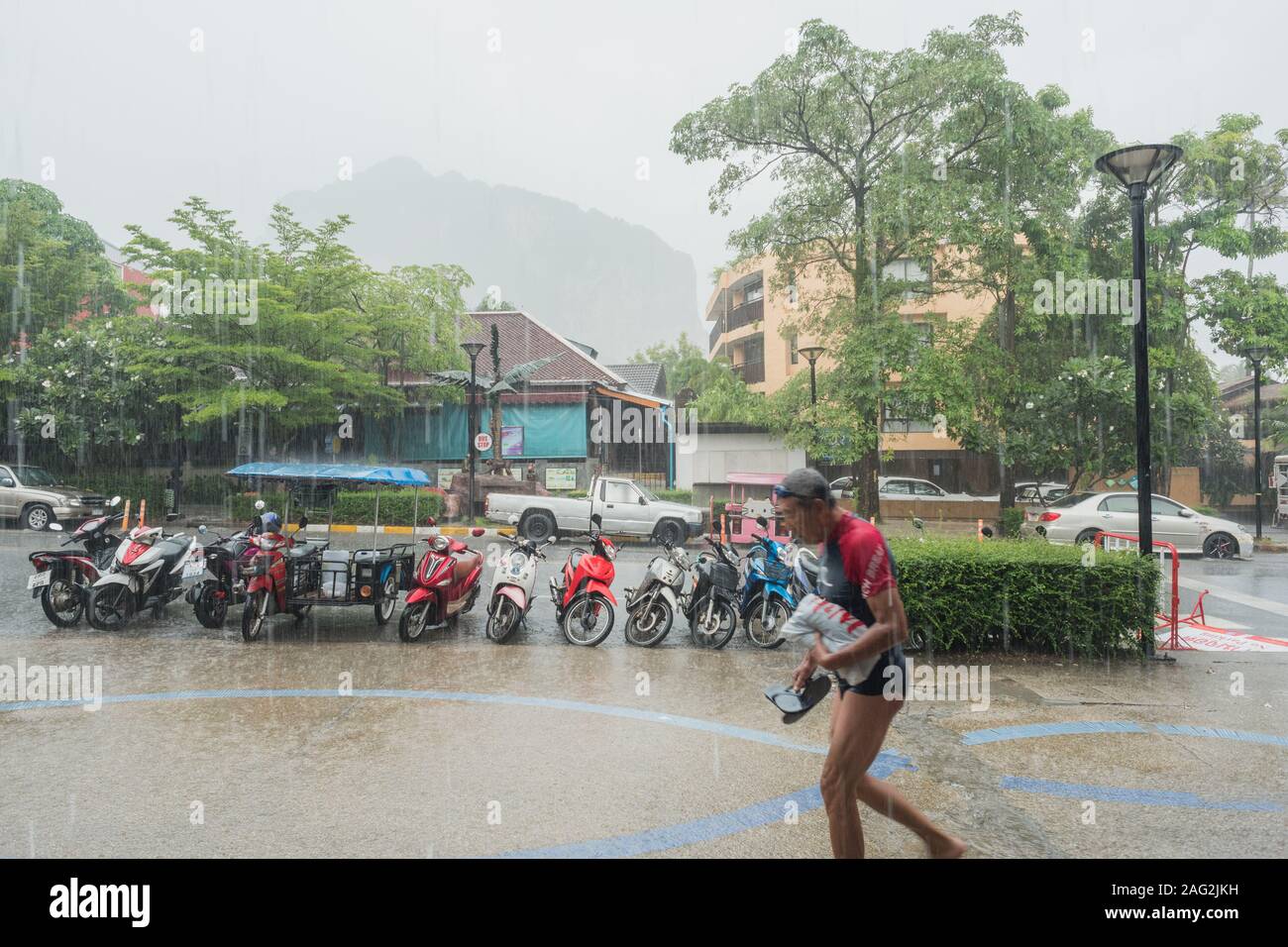 Ao Nang, Krabi/Thailand - Juli 16, 2019: ein Tourist verlangsamende durch starken Regen, der Besitz einer Plastiktüte und ein Paar Flip Flops, entlang der Hauptstraße. Stockfoto