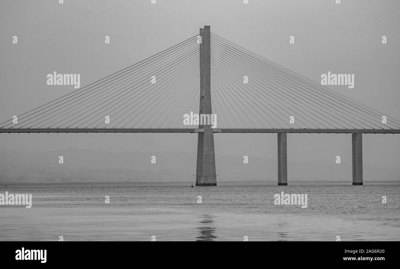 Graustufenaufnahme einer Hängebrücke in der Nähe des ruhigen Meeres Unter dem dunklen Himmel Stockfoto