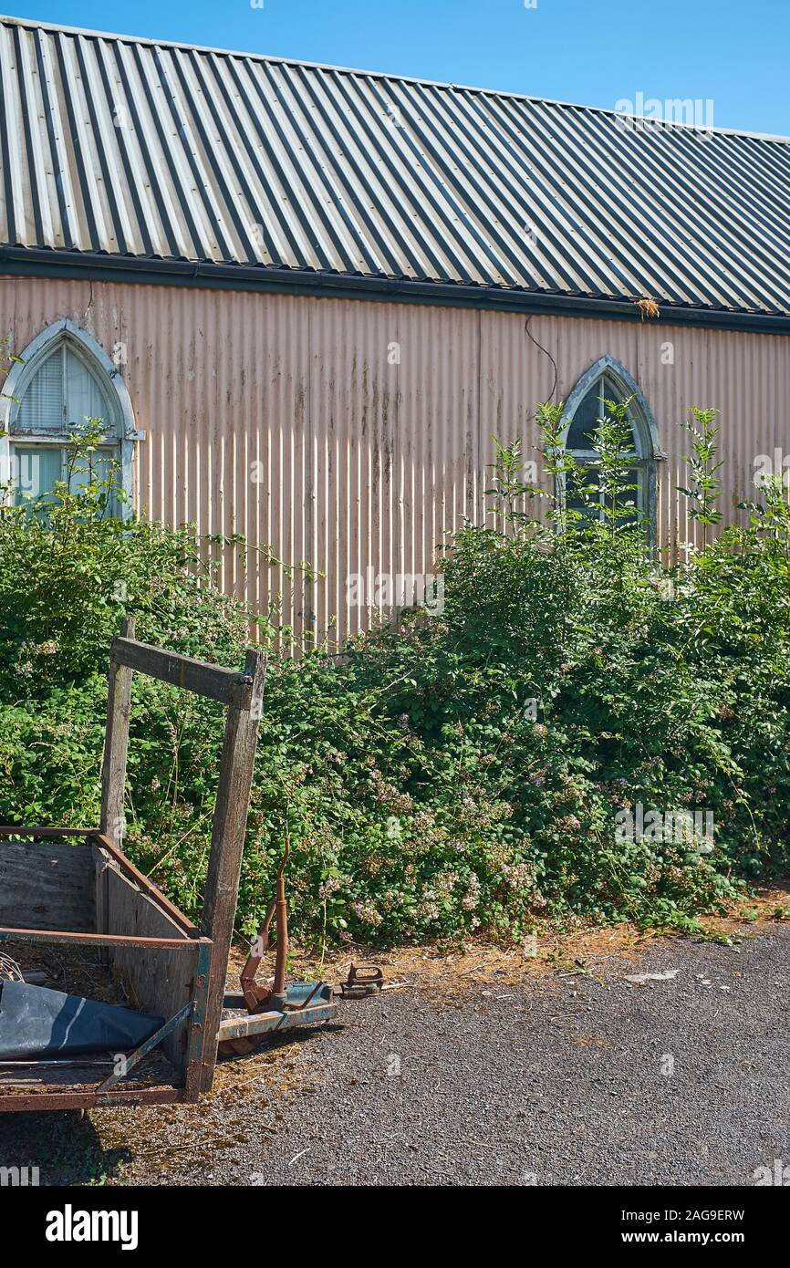 Ein altes Gebäude, das aus Wellblech gefertigt wurde und mit Spitzbogenfenstern, einer überwucherten Hecke und einem alten, abgenutzten Anhänger im Freien ausgestattet ist Stockfoto