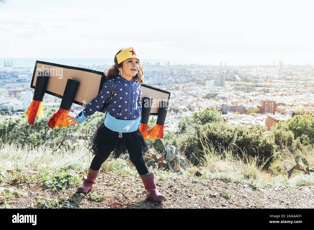 Lustige kleine Kind Mädchen fliegen als Superheld mit hausgemachten Kostüm und Karton Flugzeug Flügel vor einem Stadtbild, Phantasie und Mädchen verkleidet Stockfoto