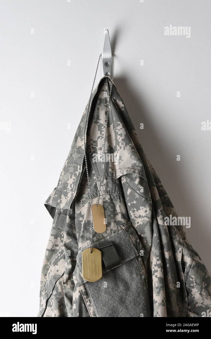 Eine Reihe von militärischen Dog Tags und Camouflage Feld Jacke hängt an einem Haken auf eine leere Wand. Stockfoto