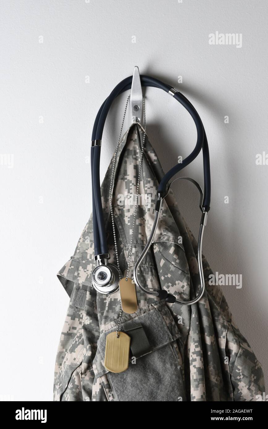 Militärische Gesundheitswesen Konzept: Eine Reihe von Dog Tags, Stethoskop und Camouflage Feld Jacke hängt an einem Haken auf eine leere Wand. Stockfoto