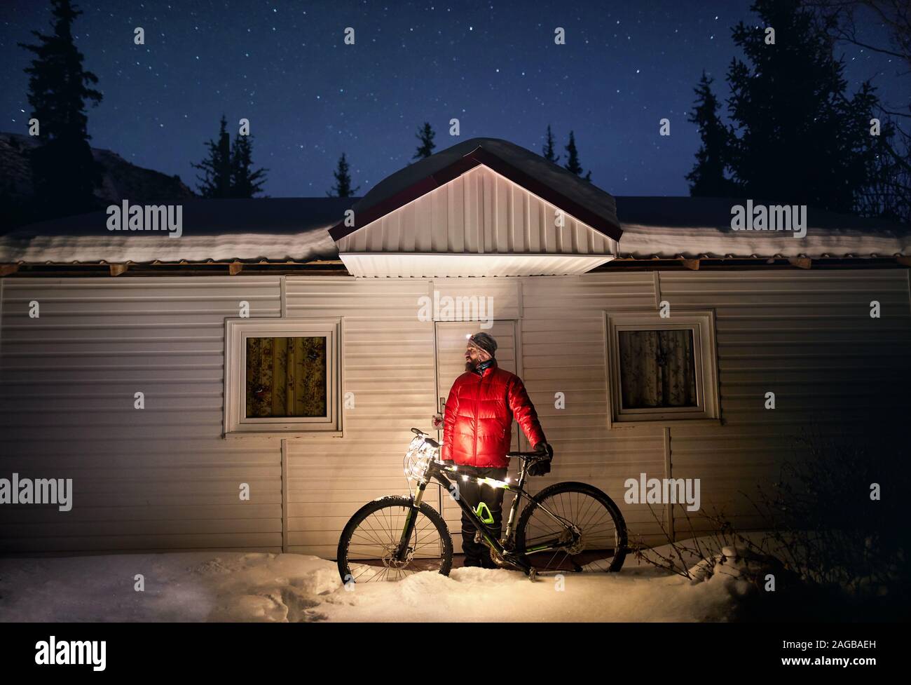 Mann im roten Mantel mit Fahrrad mit Weihnachtsbeleuchtung in der Nähe von kleines Haus im Winter verschneite Wald in die Berge unter dem Nachthimmel mit Sternen Stockfoto