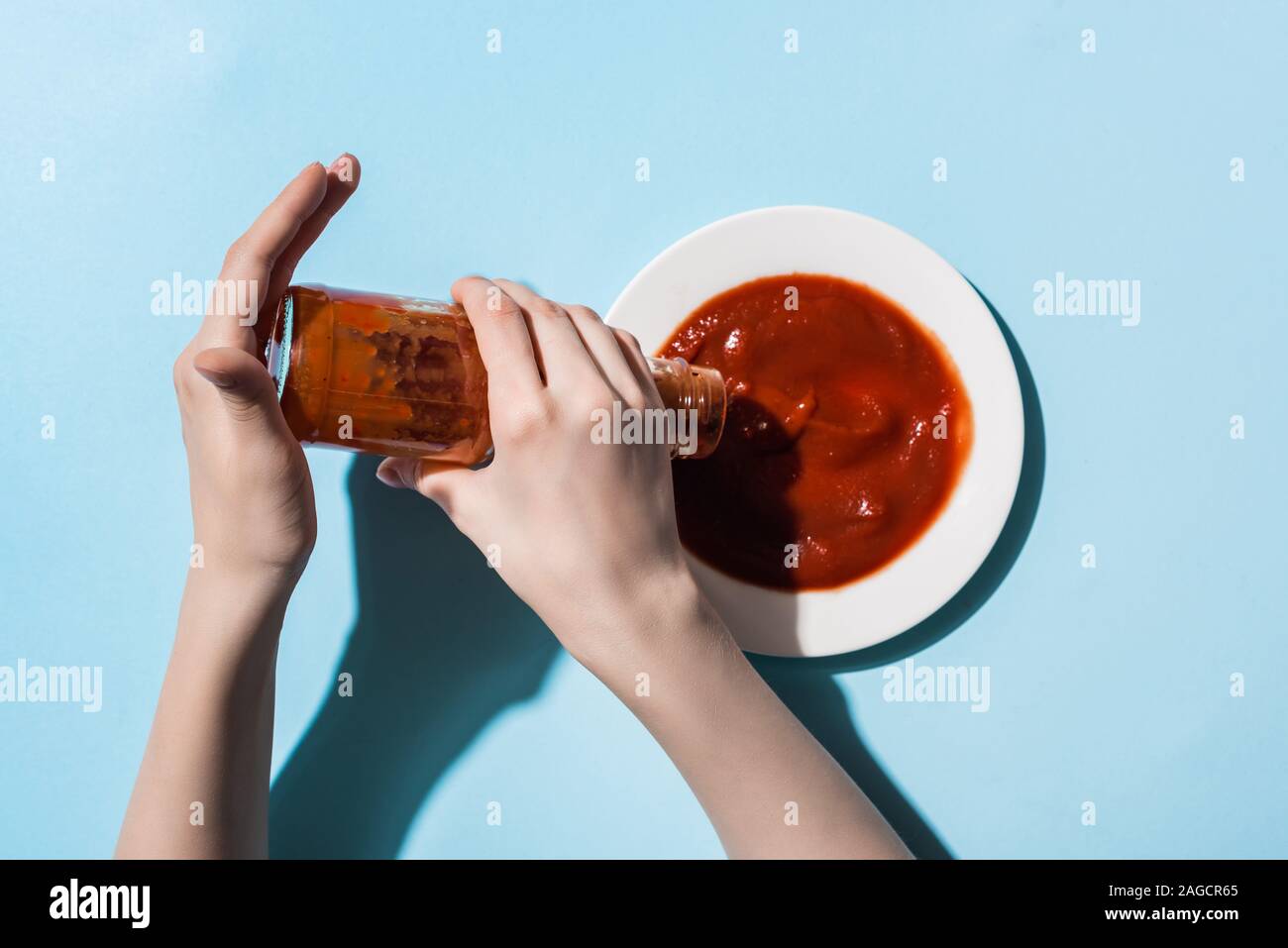 7/8-Ansicht von Frau gießen Tomatensoße aus der Flasche an der Platte auf blauem Hintergrund Stockfoto