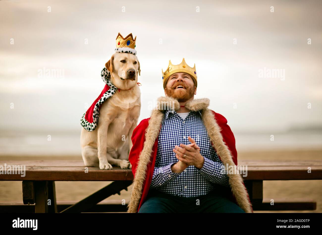 Mitte - erwachsener Mann mit Mantel und Krone und Lachen beim Sitzen auf der Bank mit seinem Hund am Strand. Stockfoto