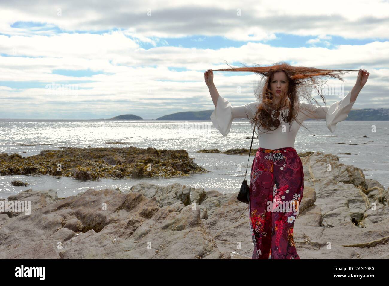 Hippie modische junge Dame mit langen braunen Haaren und floralen Schlaghosen am Meer auf Felsen unter einem cloudscape posiert, schmollen und Spielen wit Stockfoto