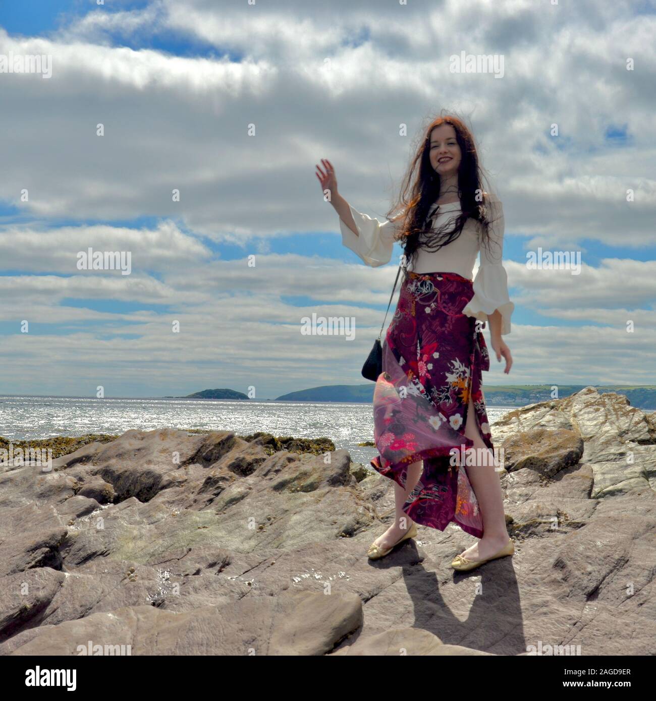 Hippie modische junge Dame mit langen braunen Haaren und floralen Schlaghosen am Meer auf Felsen unter einem cloudscape Posing, lachend mit der Hand Stockfoto