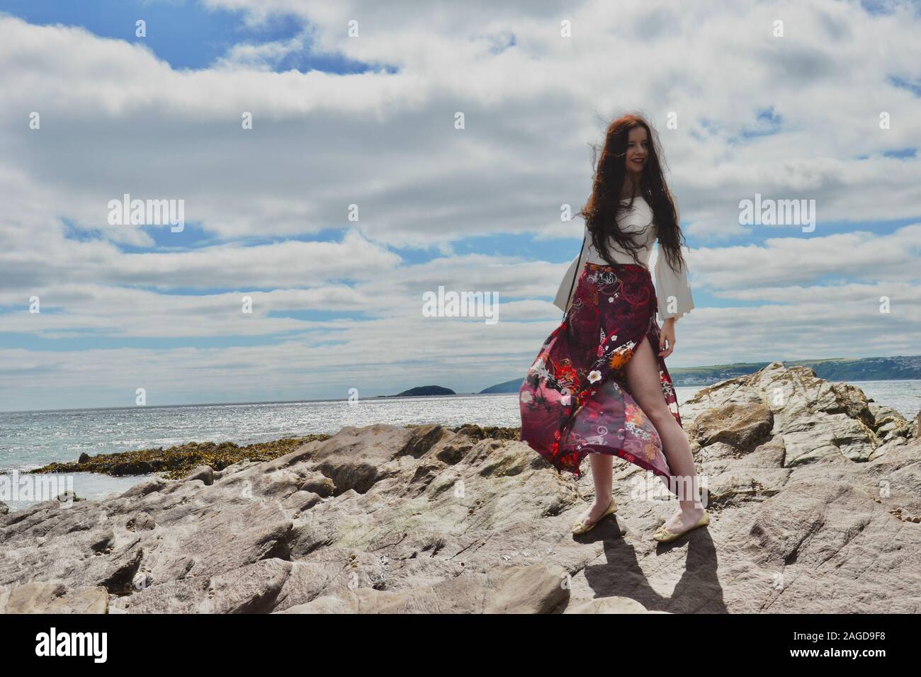 Hippie modische junge Dame mit langen braunen Haaren und floralen Schlaghosen am Meer auf Felsen unter einem cloudscape Posing Stockfoto