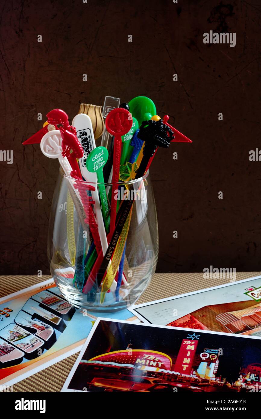 Ein klares Glas mit einer großen Anzahl an Vintage-Cocktail-Rührstäbchen in verschiedenen Farben und Formen mit alten Postkarten, die auf dem Tisch verstreut sind. Stockfoto