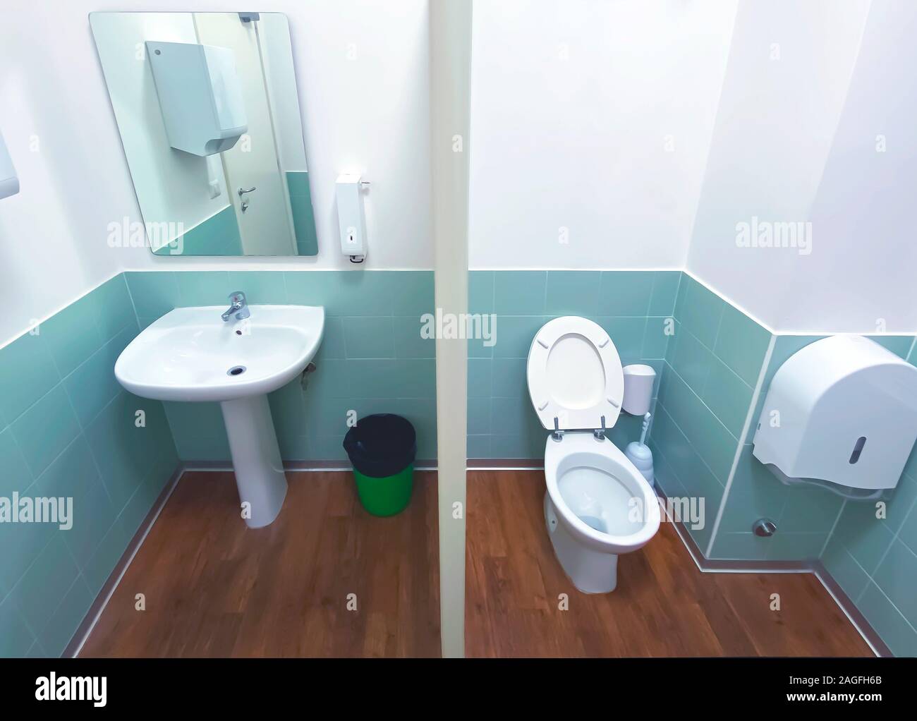 Weitwinkelaufnahme eines Wc mit dem Holzboden und gefliesten Wand mit einem Spiegel, einer grünen Korb, ein Waschbecken und die keramische Schüssel. Stockfoto