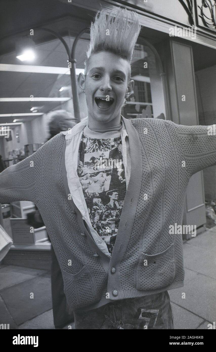 1980er Jahre, ein Bild von einem jungen Mann außerhalb eines Shop tragen eine Strickjacke über eine T-Shirt stehen und zeigt seine Whacky, unkonventionelle 'Punk' Frisur des EFR, England, UK. Wir sehen hier eine Form der "tribal Punk'Haar ähnelt einem Mohawk. Stockfoto