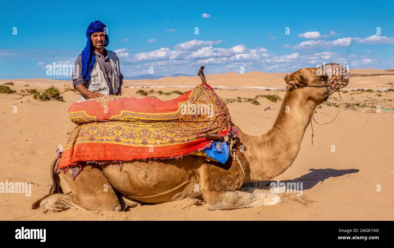 Agadir, Marokko - Okt. 21, 2015. Einen marokkanischen Mann verkauft Kamelreiten in der Wüste zu Touristen. Das Kamel ist kniend für einen Touristen zu montieren. Stockfoto