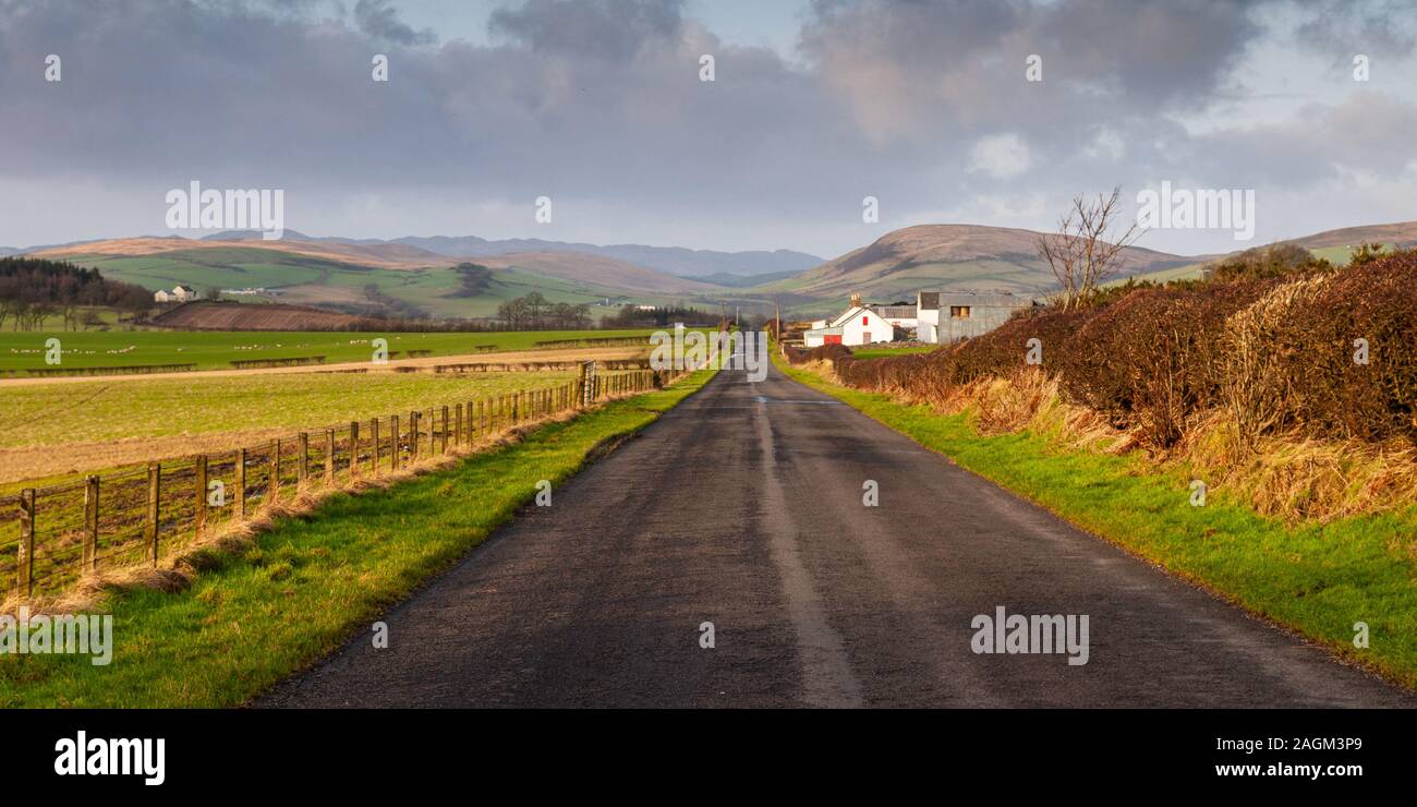 Eine lange gerade Straße verläuft, landwirtschaftliche Gebäude und Felder auf der Isle of Bute, mit hinter die Hügel und Berge der Highlands von Schottland steigt. Stockfoto