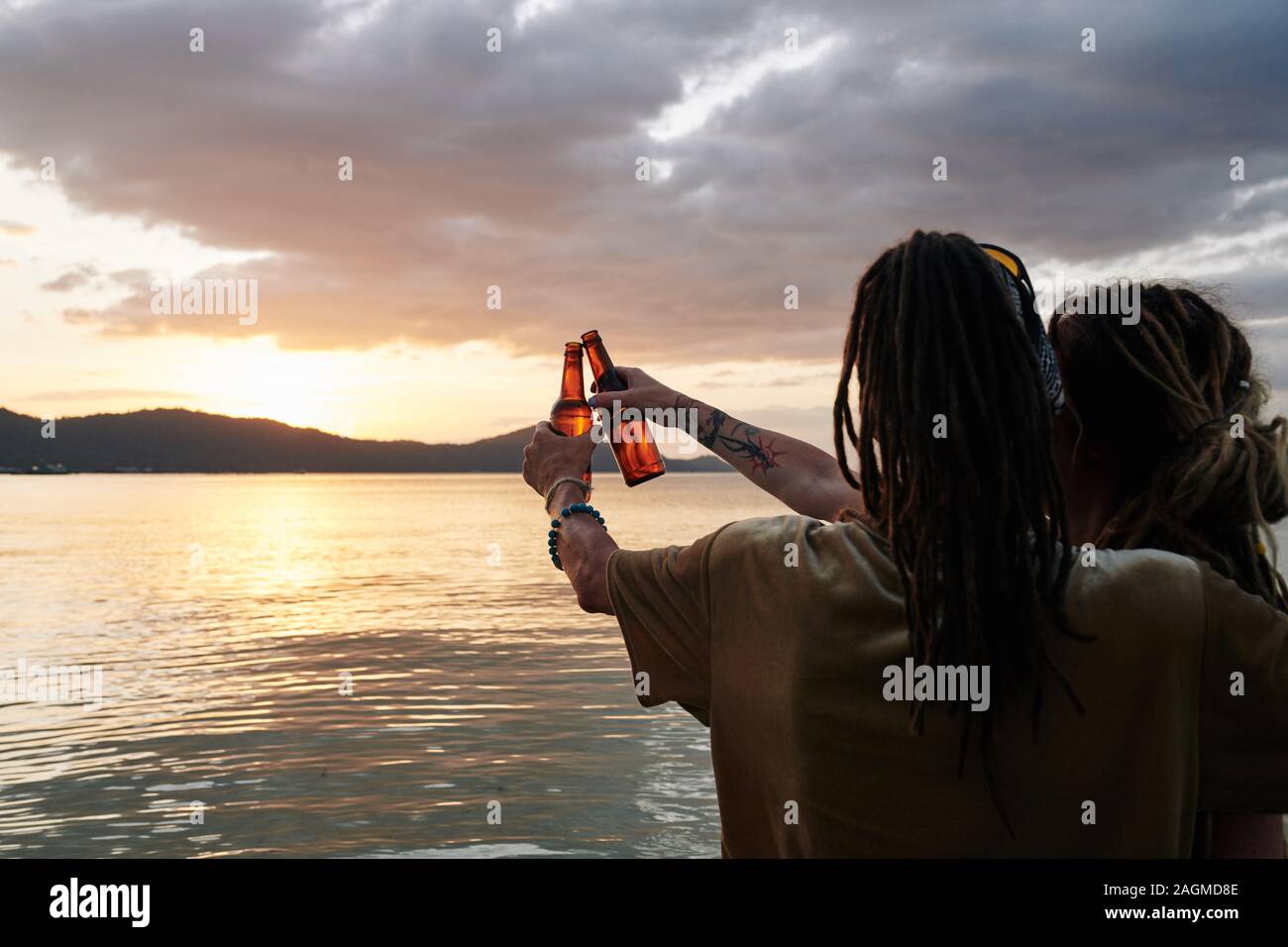 Ansicht der Rückseite des Paar genießen Sie einen wunderschönen Sonnenuntergang und Bierflaschen klirren, wenn die Ausgaben romantischen Abend am Strand. Stockfoto