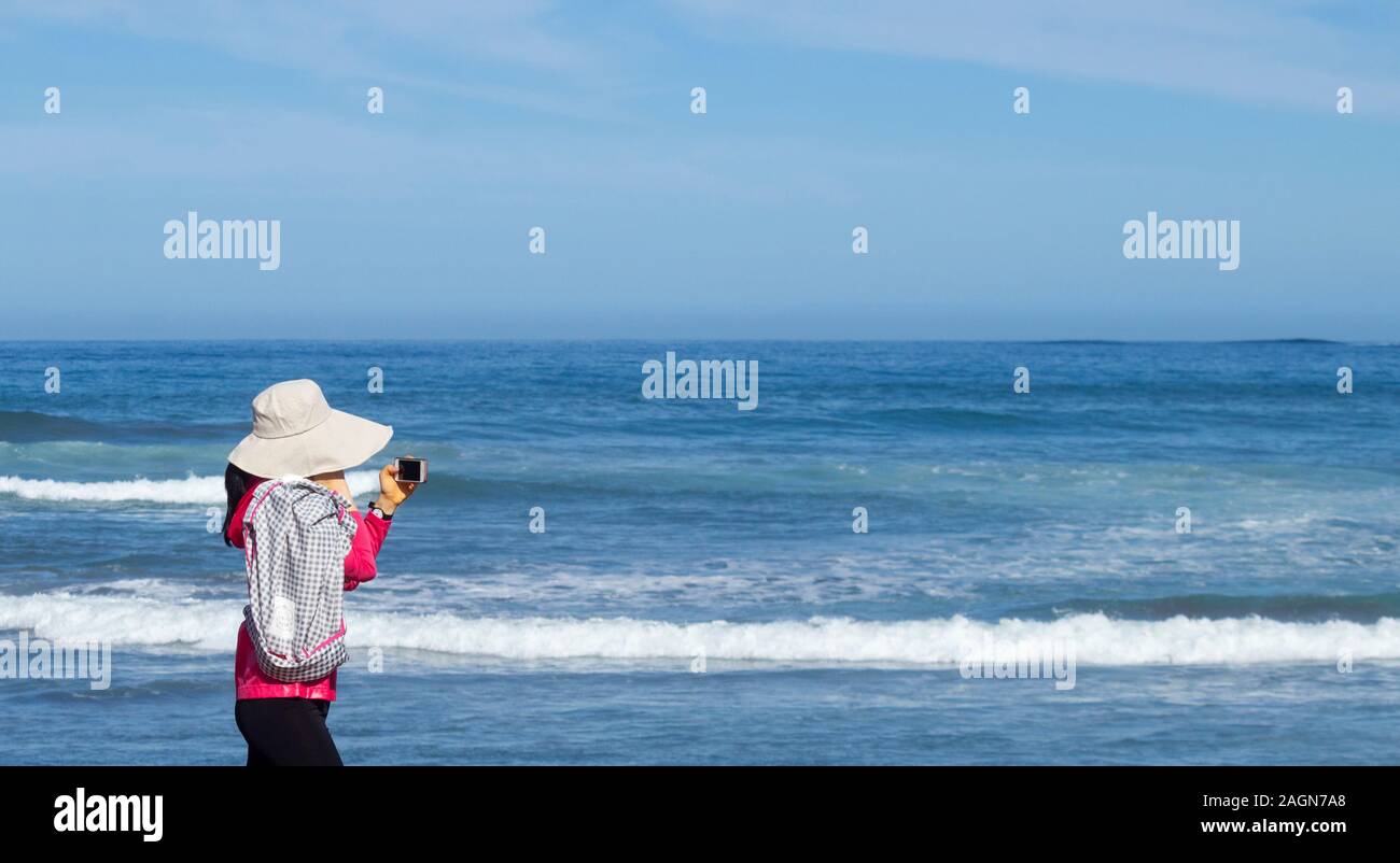 Frau mit Hut für Sonnenschutz am Handy suchen Bei einem Spaziergang am Strand. Stockfoto