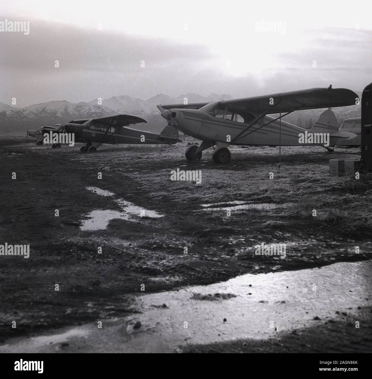 1960, historische, kleine 2-Sitzer flächenflugzeug Propeller- Flugzeugen sitzen auf unebenem Gelände in der Nähe von Flughafen Anchorage, Anchorage, Alaska, USA, mit der Chugach Mountains im Hintergrund. Stockfoto
