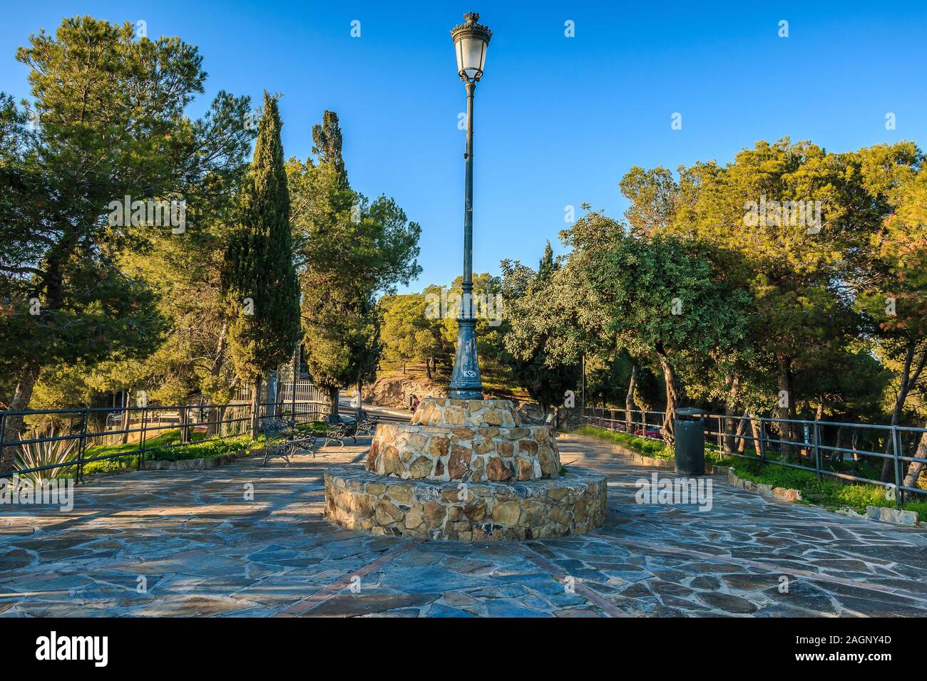 Gibralfaro in Málaga. Aussichtspunkt mit gepflastertem Fußweg und zentraler Straßenleuchte am sonnigen Tag umgeben von Nadelbäumen und Becken mit blauem Himmel Stockfoto