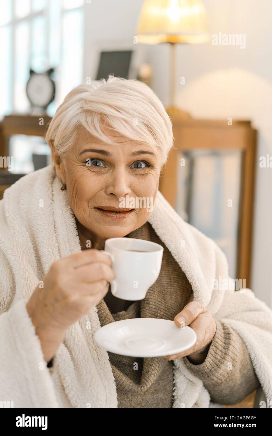Gut aussehender Älterer Frau eingehüllt in ein warmes, weißes Plaid ist ein zu Hause. Sie sitzt auf dem gemütlichen Sofa und halten Kaffee Becher in der Hand. Die Stockfoto