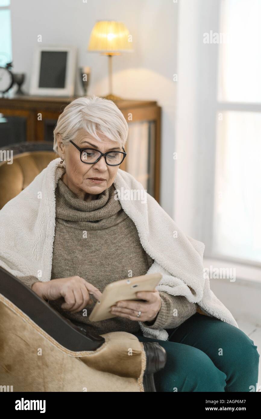 Thougtful alte Dame in Gläsern ist entspannend auf Ihre komfortable Wohnung. Sie sitzt auf dem Leder Sessel und mit Ihrem Tablet-PC. Die Frau ist Gewickelt Stockfoto