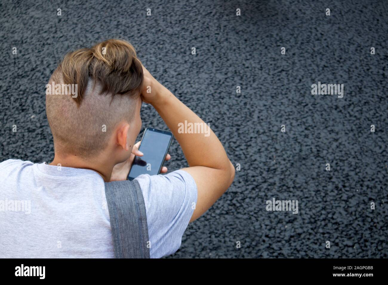Rückseite der Kopf rasiert mit Pferdeschwanz junger Mann mit Smartphone gegen grauen Asphalt Stockfoto