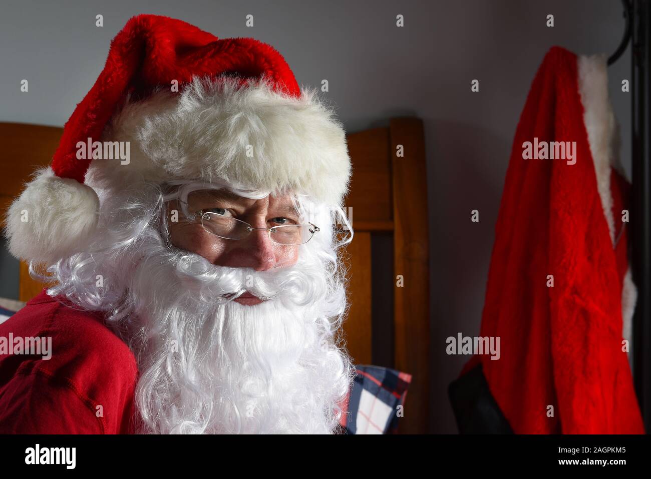 Santa Claus saß auf seinem Bett in seinem Long Johns mit seinem roten Anzug hängt an einem Haken im Hintergrund. Stockfoto