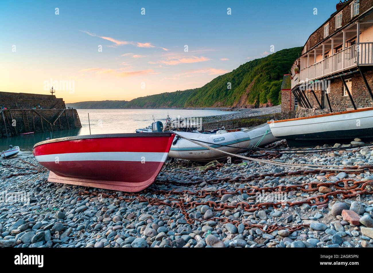 Rot-weißes Boot, das am Kiesstrand des Fischerdorfes Clovelly festgemacht ist, mit Gebäuden und Klippen und dem Hafen im Hintergrund, Südwesten. Stockfoto