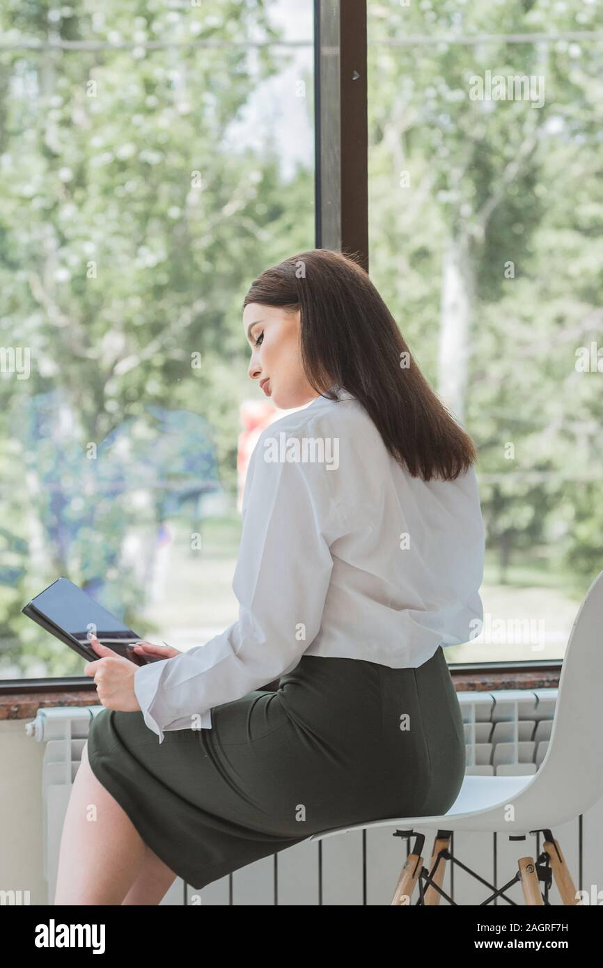 Junge schöne Frau in einem Anzug sitzt vom Amt Fenster in ihren Händen einen Tablet-PC sieht Sie in der Reflexion wie in einem Spiegel. Stockfoto