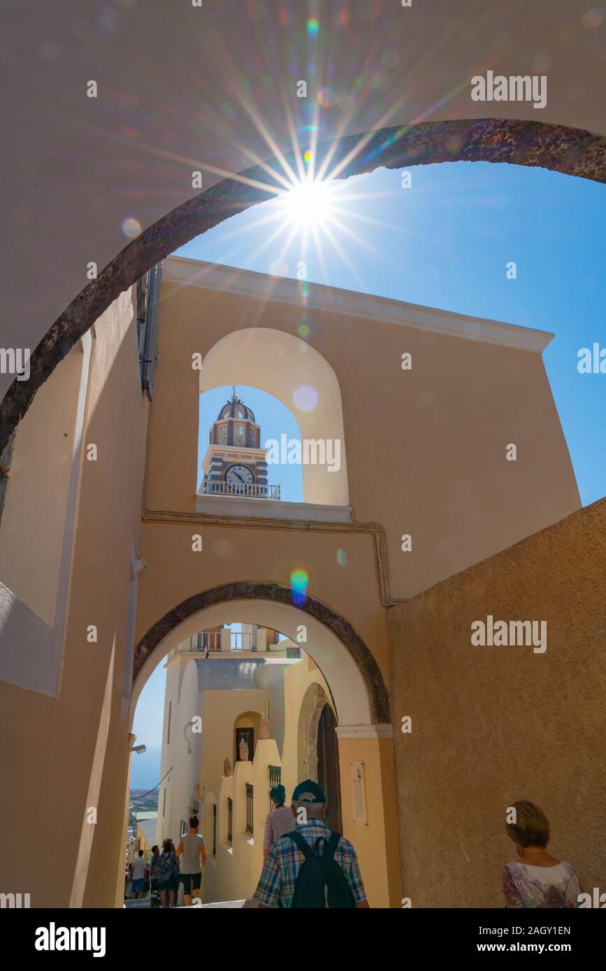 Santorini Griechenland - 8. August 2019; Sun blasts durch unter einem Bogen im Line up der architektonischen Merkmale der Bögen, Glockenturm und Uhr in Fira, der als Stockfoto