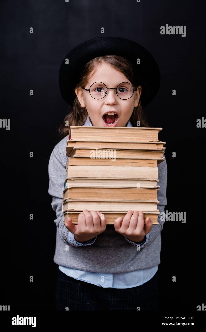 Aufgeregt elementare Schülerin in Hut und Brille tragen Stapel Bücher auf schwarzem Hintergrund isoliert Stockfoto