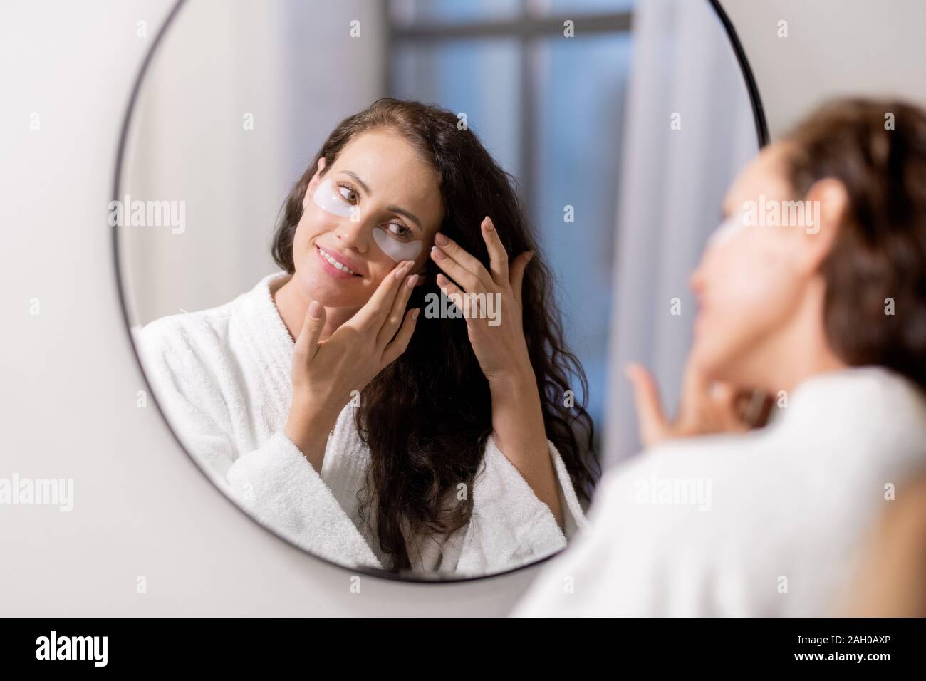 Glückliche junge Frau im weißen Bademantel putting undereye Patches auf dem Gesicht Stockfoto