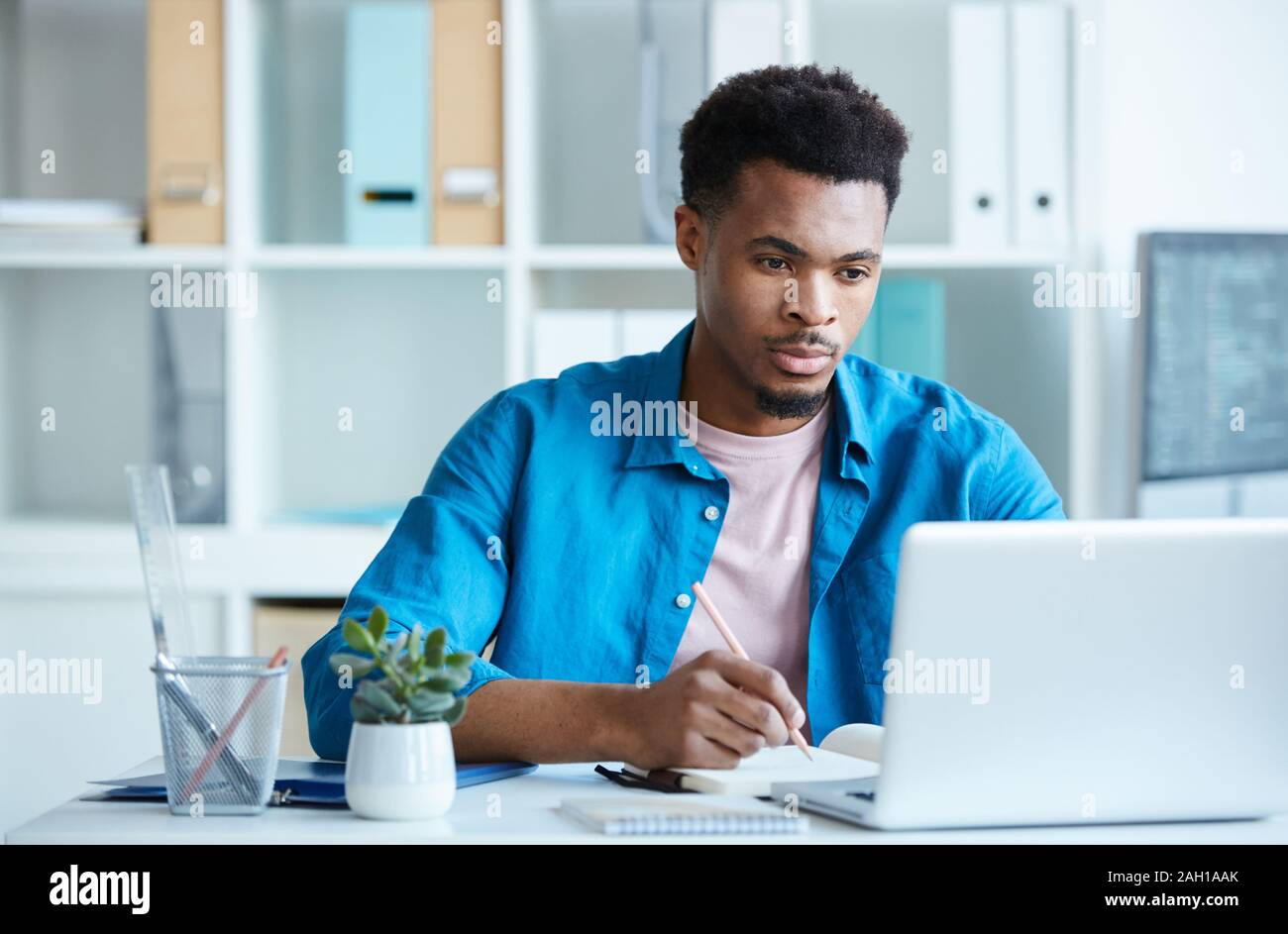 Schweren Afrikanischen jungen Mann in Freizeitkleidung am Tisch sitzen mit Blick auf die Überwachung von Laptop und Notizen im Büro Stockfoto