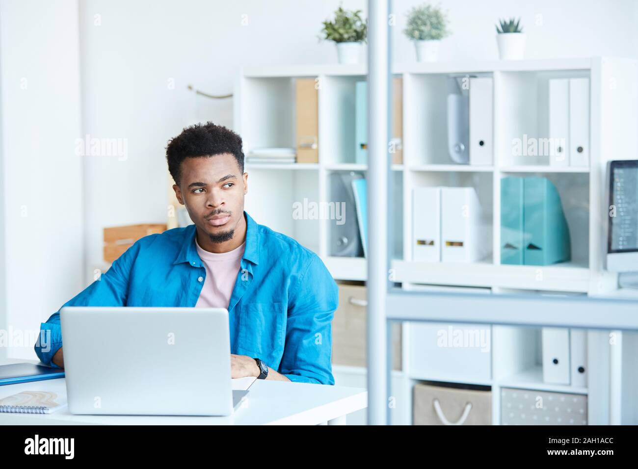 Afrikanische nachdenklicher Mann in Freizeitkleidung sitzt am Tisch vor Laptop und Denken über etwas im Büro Stockfoto