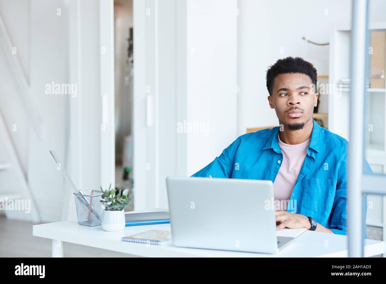 Afrikanischer junge Unternehmer in blau casual Shirt an seinem Arbeitsplatz sitzt mit Laptop mit nachdenklichen Blick Stockfoto
