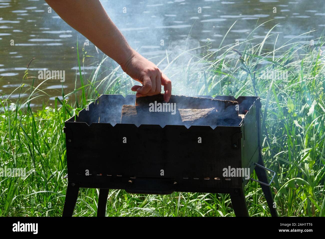 Grill mit Kraftstoff und Holzkohle. Hand ist das Hinzufügen von Holz in das Feuer. Grill in der Nähe des Flusses. Sun. Stockfoto