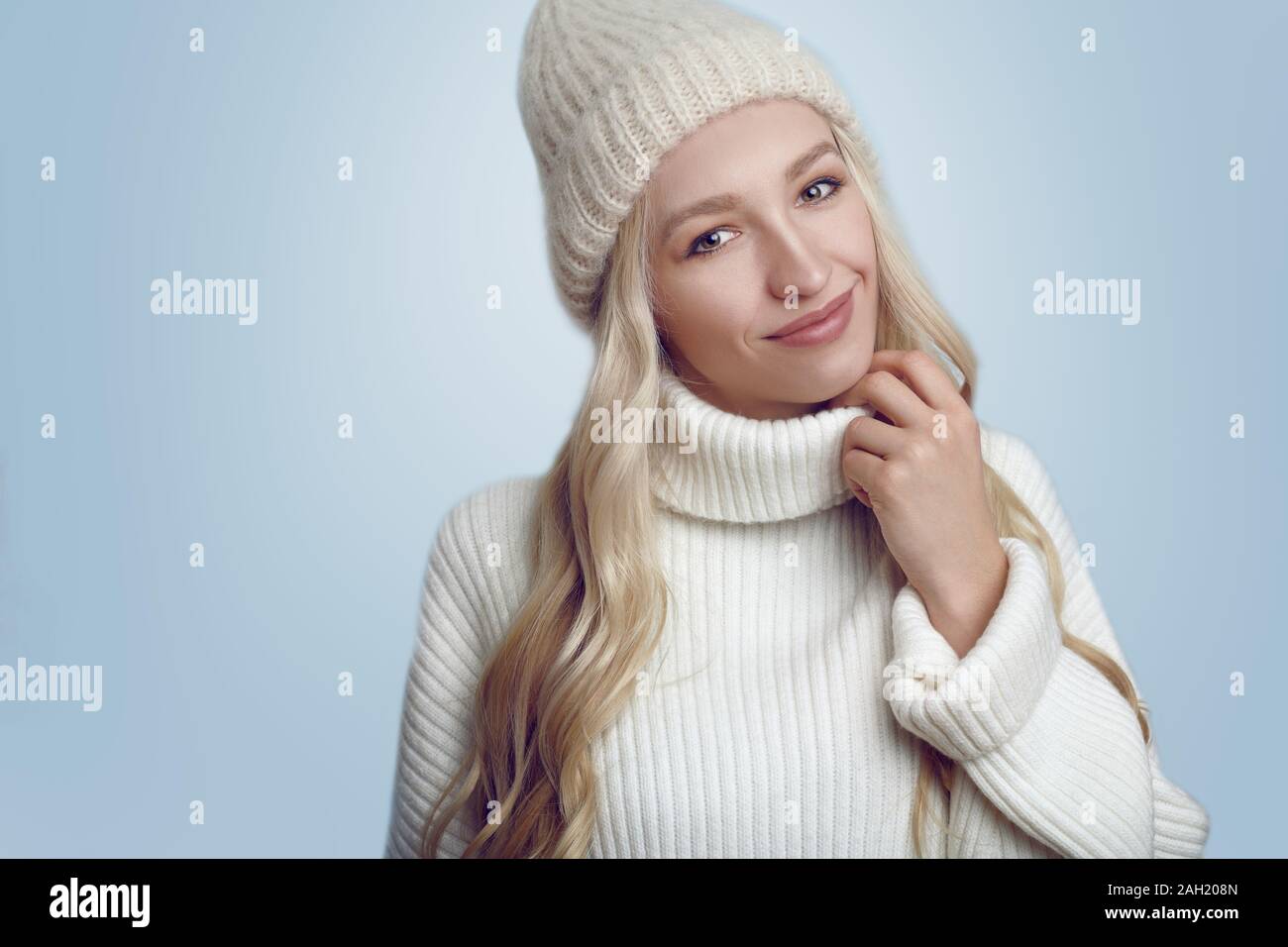 Schöne junge blonde Frau in Weiß gestrickte Pullover und Hut lächelnd und mit Blick auf die Kamera. Close-up front Portrait gegen Uni hellblau backgroun Stockfoto