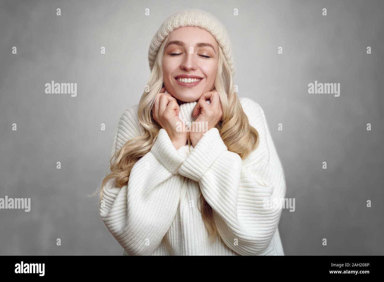 Junge blonde Frau, die ihre weißen Strickpullover genießen, gegen grauen Hintergrund stehend, mit den Händen an den Hals und lächelnd mit geschlossenen Augen Stockfoto