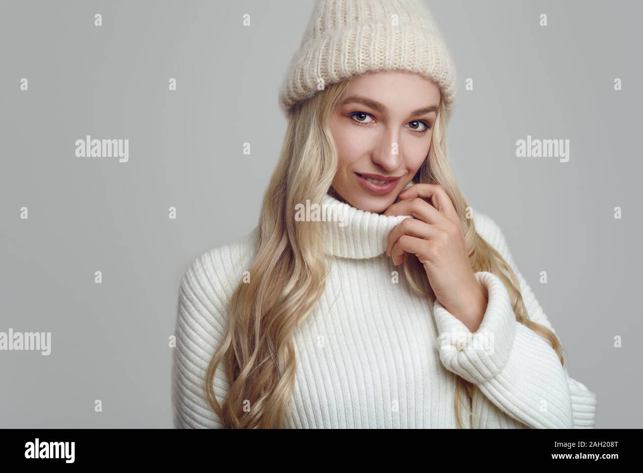 Schöne junge blonde Frau in Weiß gestrickte Pullover und Hut lächelnd und mit Blick auf die Kamera. Close-up front Portrait gegen Uni hellblau backgroun Stockfoto