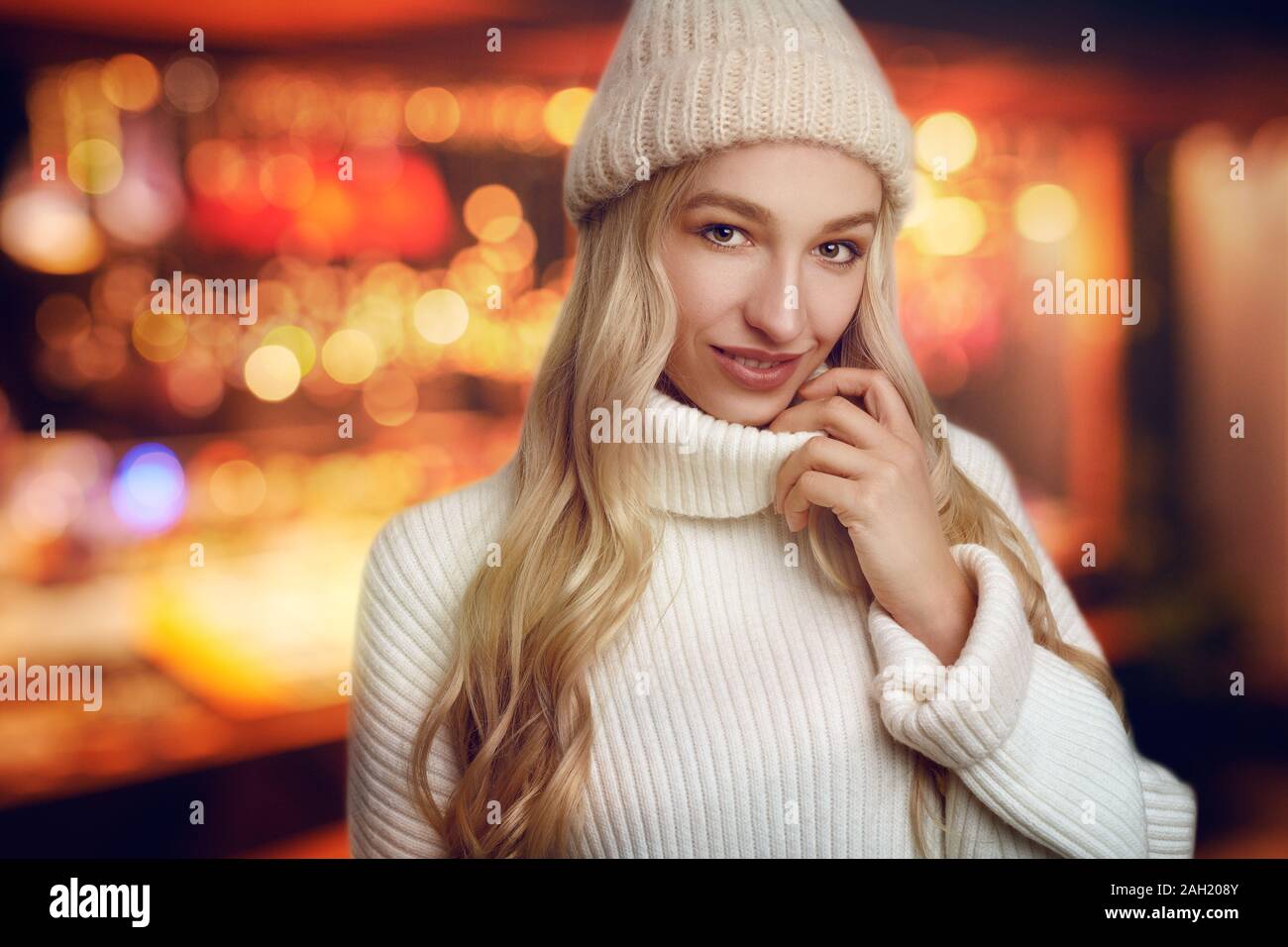 Schöne junge blonde Frau in Weiß gestrickte Pullover und Winter Hut, von der Kamera schauen und lächeln, stehend gegen warme Gelb leuchten mit b verschwommen Stockfoto