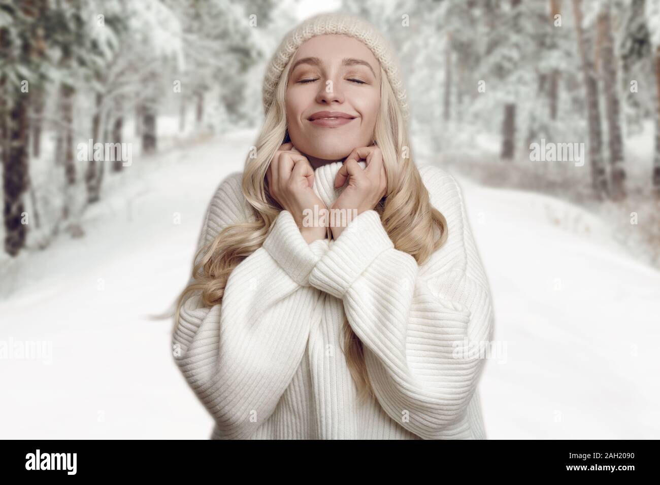 Junge blonde Frau, die ihre weißen Strickpullover genießen, gegen Winter wald landschaft Hintergrund stehend, mit den Händen an den Hals und mit dem Lächeln Stockfoto
